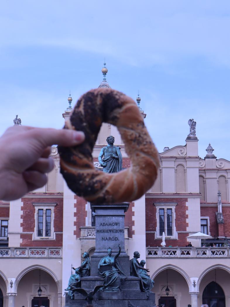 Eine Hand hält einen traditionellen Krakauer Brezel (Obwarzanek) in die Kamera, sodass er das Denkmal des polnischen Dichters Adam Mickiewicz auf dem Hauptmarkt von Krakau einrahmt. Im Hintergrund ist die Fassade der historischen Tuchhallen (Sukiennice) m