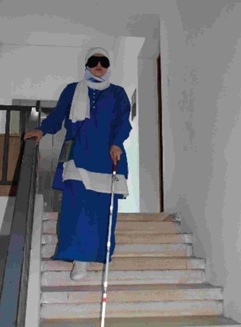 2.	Eine Frau geht mit Langstock unter der Augenbinde eine Treppe hinunter. Sie trägt Kleid und Hijab in weiß und blau. 
