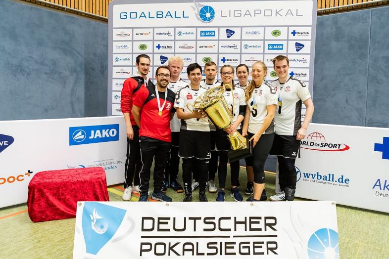 Foto der Goalballmannschaft mit kindergroßem Pokal hinter einem kniehohen Schild mit der Aufschrift "Deutscher Pokalsieger".