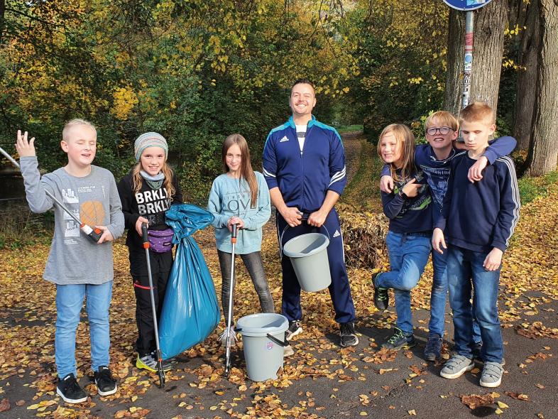 6 Schülerinnen und Schüler der 5a posieren mit Lehrer und der Müllsammel-Ausrüstung an einer Wegkreuzung im Grünen.