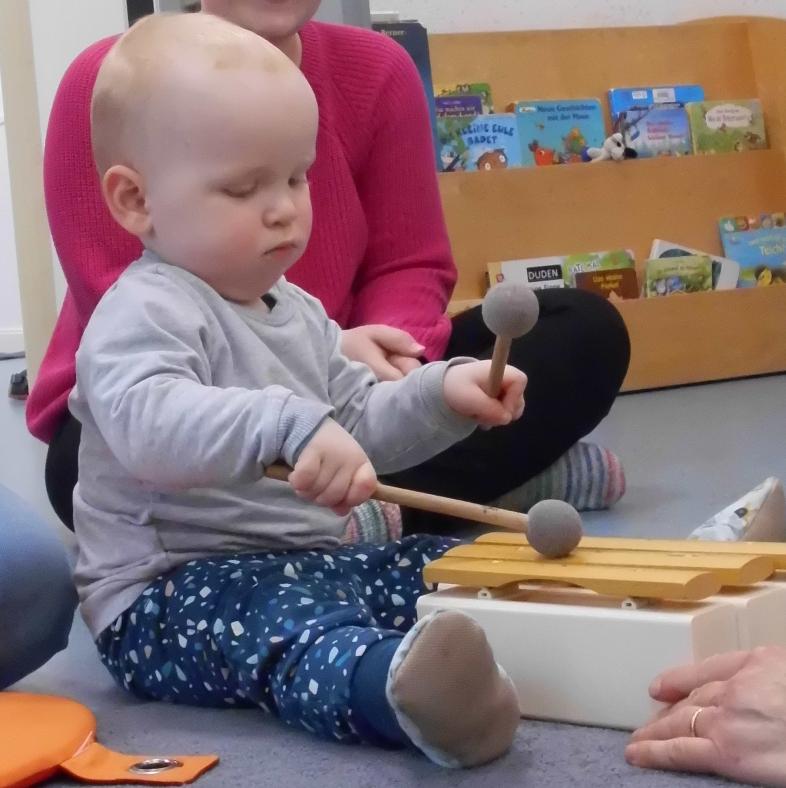 Ein Kleinkind spielt mit zwei Holzschlägeln ein Xylophon. In Hintergrund ist eine erwachsene Person zu erkennen.