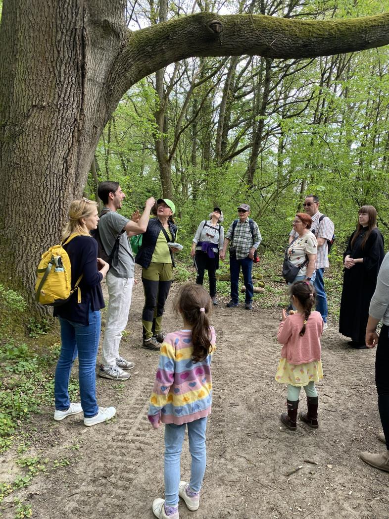 Eine Gruppe von Erwachsenen und zwei Kindern steht auf einem Waldweg unter einem großen Baum. Eine Person in der Mitte scheint der Gruppe etwas zu erklären.