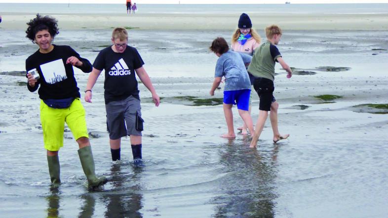 Fünf Jugendliche stapfen vergnügt barfuß oder in Gummistiefeln durch eine seichte Stelle am Nordseestrand.