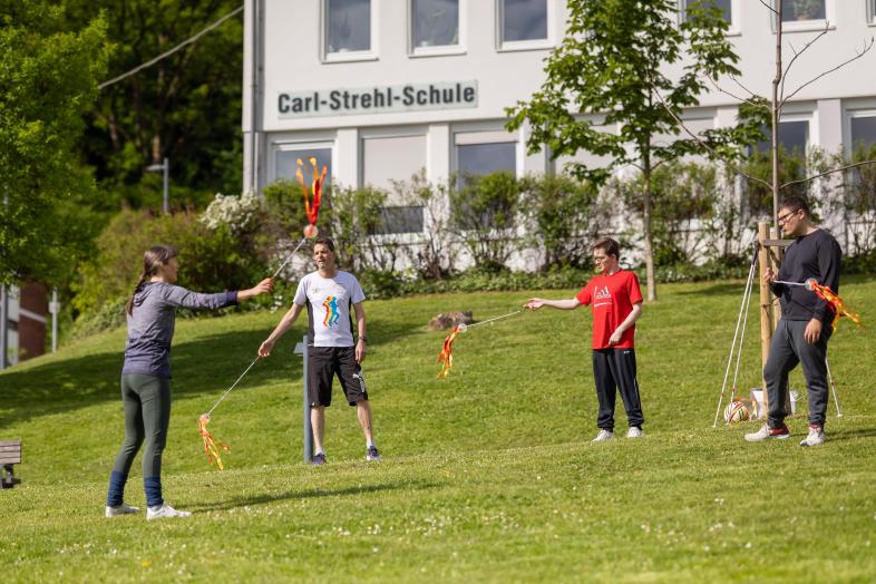 Eine Frau und ein Mann in Sportkleidung spielen auf einer Wiese auf dem blistaCampus und schwingen lange Stäbe mit bunten Bändern an den Enden.