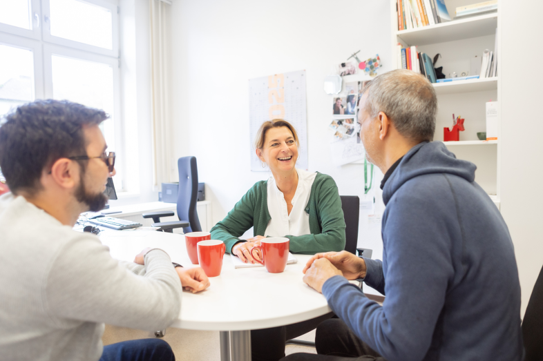Unsere Beraterin sitzt mit zwei klienten am Tisch. Sie unterhalten sich.