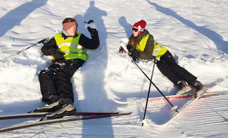 Zwei Schüler*innen liegen in Langlauf-Ausrüstung im Schnee.
