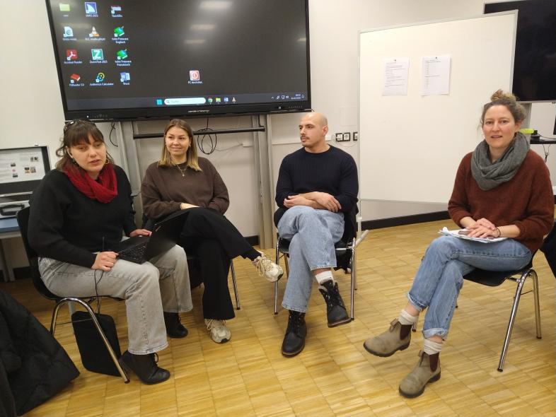 Sara und Nikolaos Rizidis sitzen mit zwei Kolleginnen vom Psychologischen Dienst auf Stühlen in einem Klassenraum. 