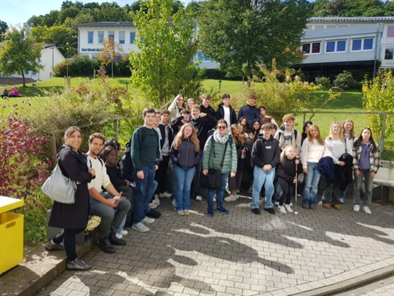 Die gruppe aus blista-Schüler*innen und spanischen Besucher*innen, ca. 30 Personen sitzen und stehen auf und vor einer Bank auf dem blistaCampus.