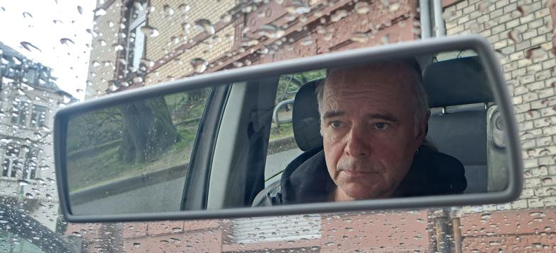 Herr W ist im Rückspiegel eines Autos zu sehen, auf dessen Windschutzscheibe Regentropfen die Sicht auf ein Haus verdecken.