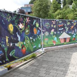 Den rund 15 Meter langen Bauzaun für die Erweiterung der Montessori-Schule auf dem Gelände der Deutschen Blindenstudienanstalt ziert die Reproduktion des Gemäldes „Sekundenschlaf“ der Marburger Künstlerin Uli Krappen. Ein farbenfrohe Traummotiv 