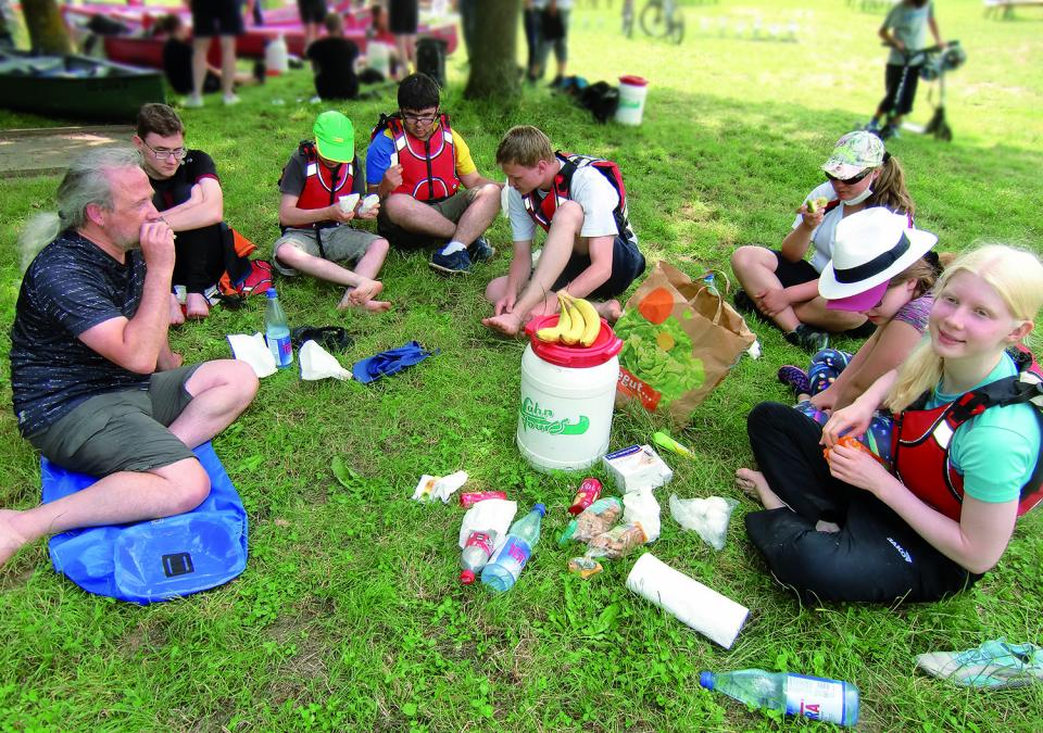 Bild 1 von 6: Foto einer Picknick-Szene auf der Wiese mit der wasserdichten Tonne in der Mitte und den Paddler*innen außenrum.