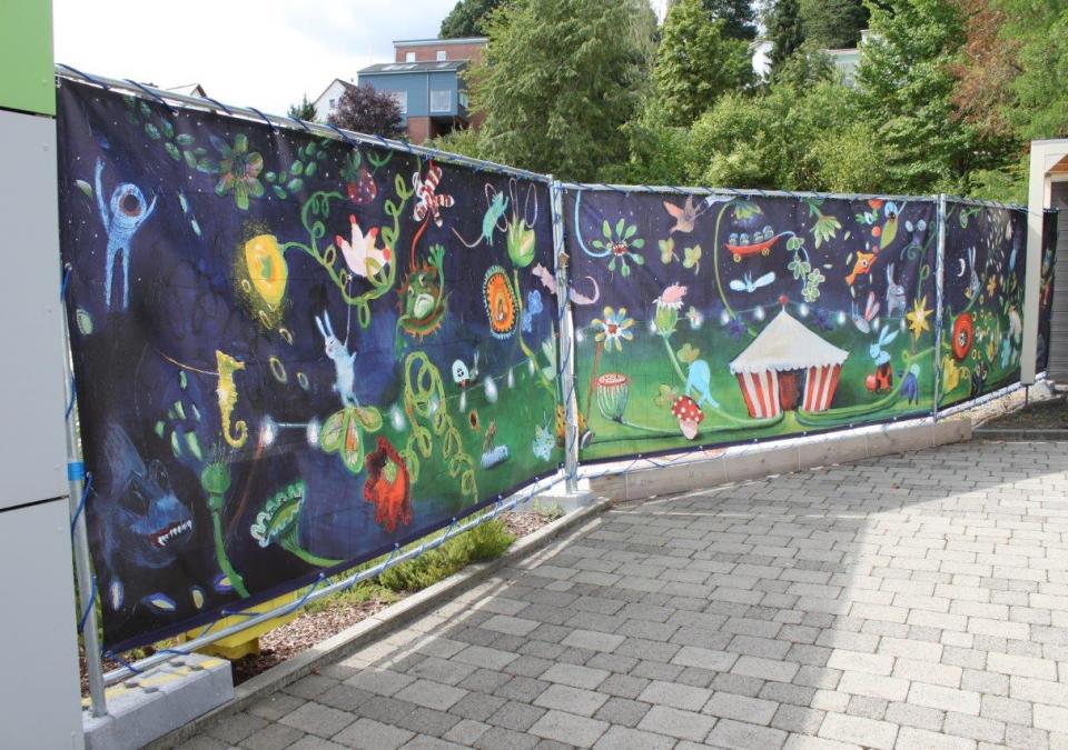 Bild 1 von 12: Den rund 15 Meter langen Bauzaun für die Erweiterung der Montessori-Schule auf dem Gelände der Deutschen Blindenstudienanstalt ziert die Reproduktion des Gemäldes „Sekundenschlaf“ der Marburger Künstlerin Uli Krappen. Ein farbenfrohe Traummotiv 