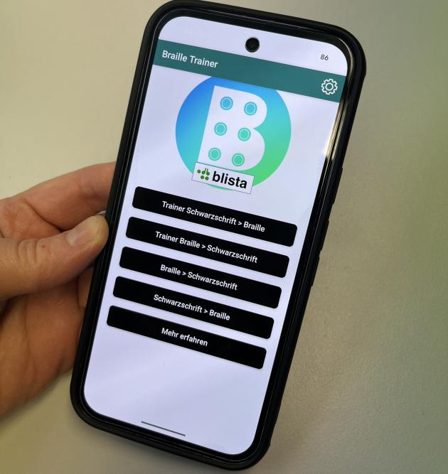 Eine Hand hält ein Smartphone, auf dem die Braille-Trainer App zu sehen ist.