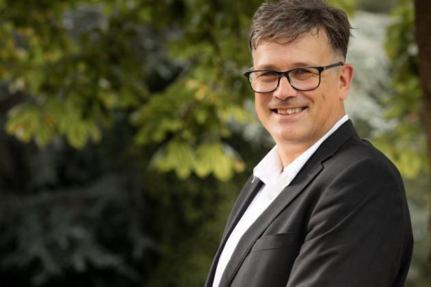 Otfid Altfeld steht im Anzug auf dem blistaCampus. Er ist groß, hat kurze Haare und trägt eine Brille. Er steht seitlich der Kamera zugewandt und lächelt-