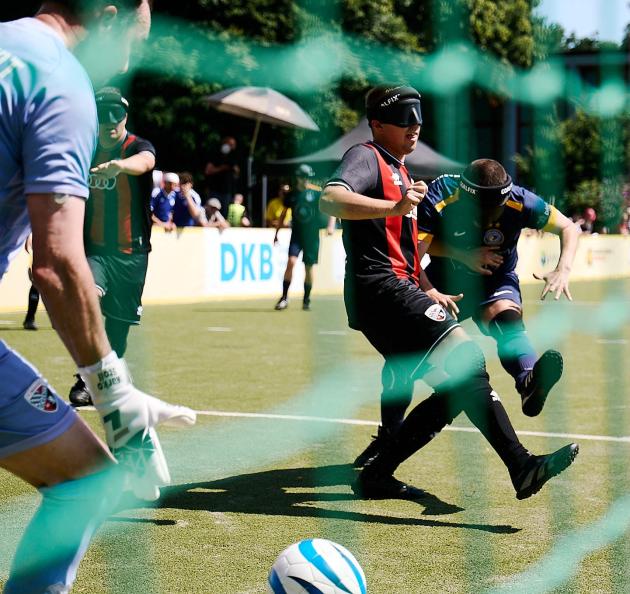 Blick durch ein Tornetz im Blindenfußball, Zwei Spieler im Duell, einer schießt den Ball in Richtung Tor.