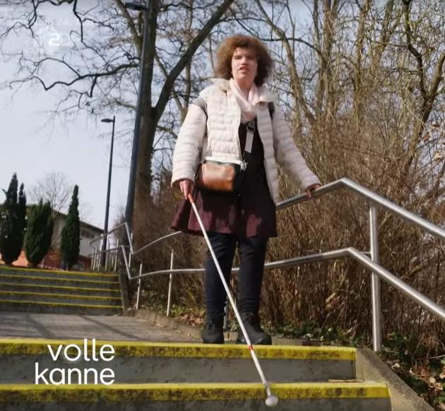 Screenshot aus dem TV-Beitrag: Eine junge Frau mit Langstock geht eine Treppe auf dem blistaCampus hinunter,