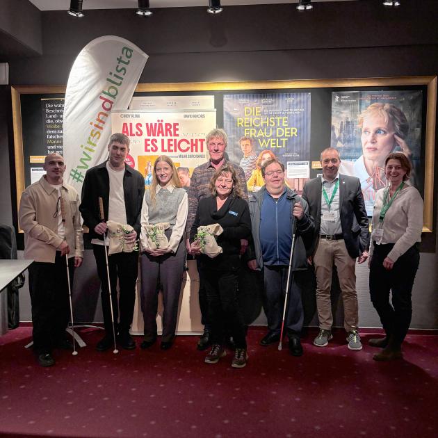 Eine Gruppe von acht Personen steht bei der Vorpremiere des Films „Als wäre es leicht“ vor Kinoplakaten und lächelt in die Kamera, einige halten Blindenstöcke oder kleine Geschenke. Im Hintergrund sind Filmplakate und ein Banner zu sehen.